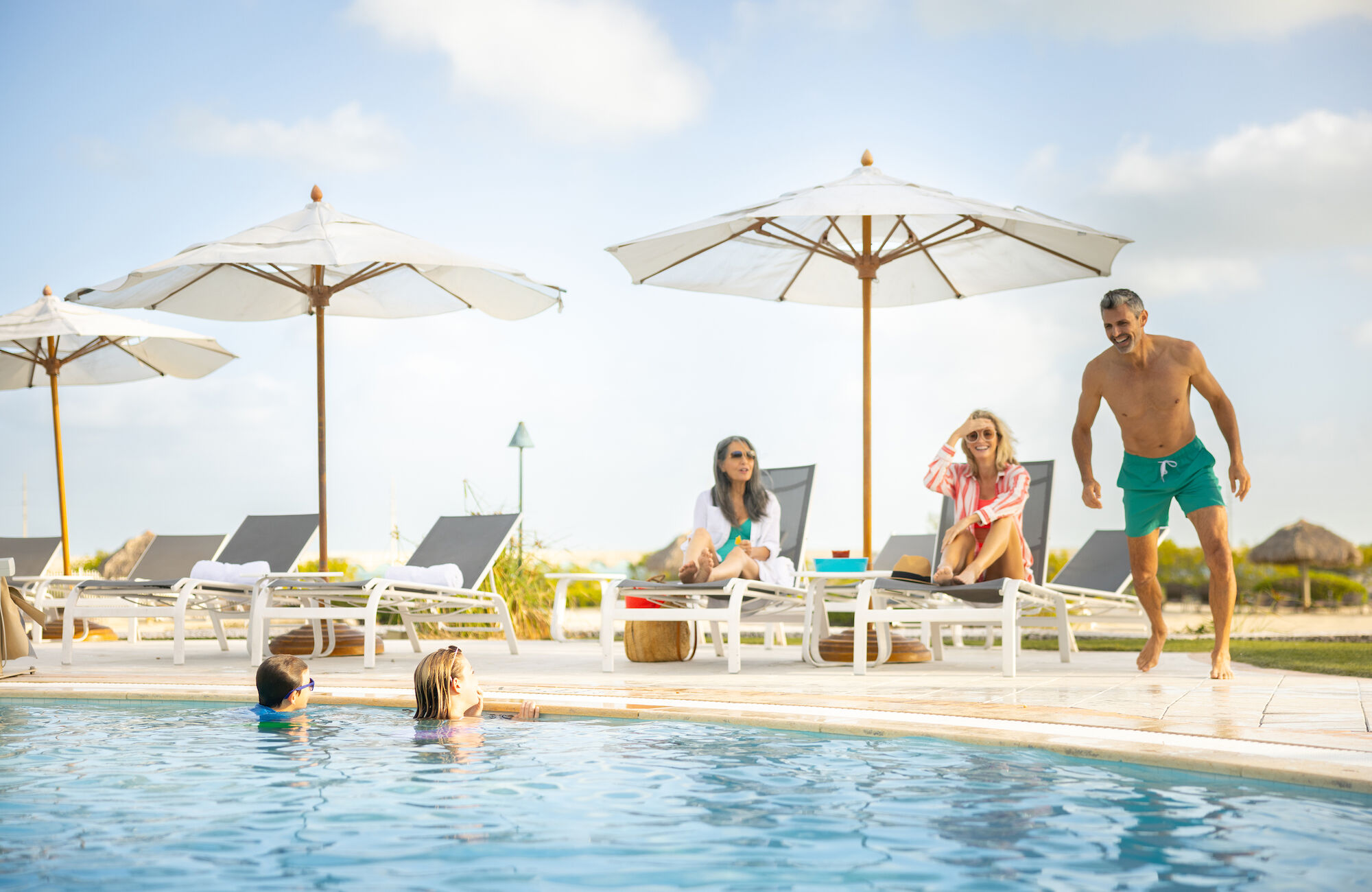 People relaxing by a pool, some lounging on chairs under umbrellas and others swimming, all enjoying a sunny day.
