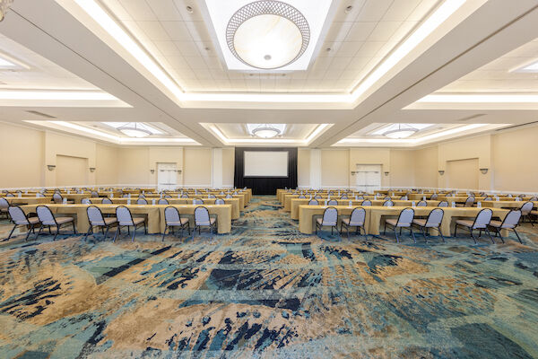 A spacious, well-lit conference room with rows of tables, chairs, water bottles, and a projector screen is visible.
