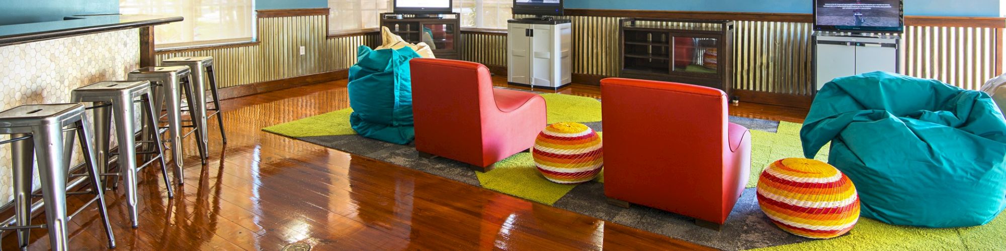 The image depicts a game or lounge room with bean bags, armchairs, multiple TVs, bar stools, and a polished wooden floor, creating a cozy atmosphere.