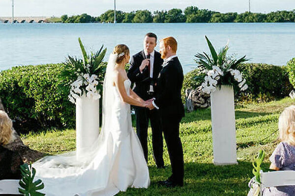 A wedding ceremony is taking place outdoors by a lake, with guests seated on white chairs under palm trees as a couple stands at the altar.