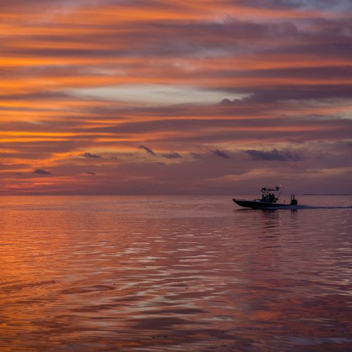 A small boat travels across calm waters under a vibrant orange and purple sunset sky with scattered clouds, creating a serene and picturesque scene.