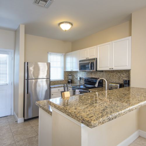 A modern kitchen with granite countertops, white cabinets, stainless steel appliances, and beige tile flooring.