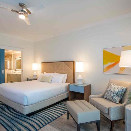 A modern bedroom with a bed, blue door, armchair, lamps, abstract art, and an attached bathroom in a well-lit and neatly arranged space.