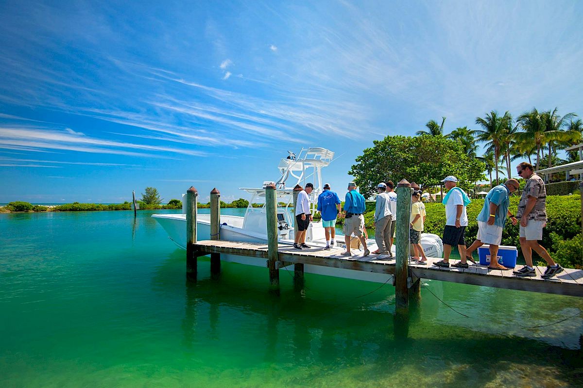 A group of people is standing on a dock by a vibrant turquoise body of water, preparing to board a boat under a clear, sunny sky with scattered clouds.