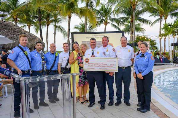 A group of firefighters poses with a large ceremonial check in an outdoor setting with palm trees and a pool.