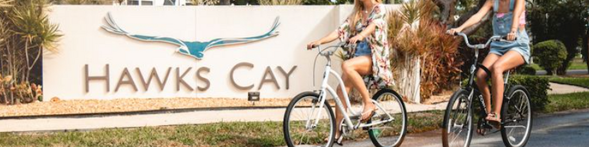 Two people are biking past a sign that reads "Hawks Cay," with palm trees and a building in the background.