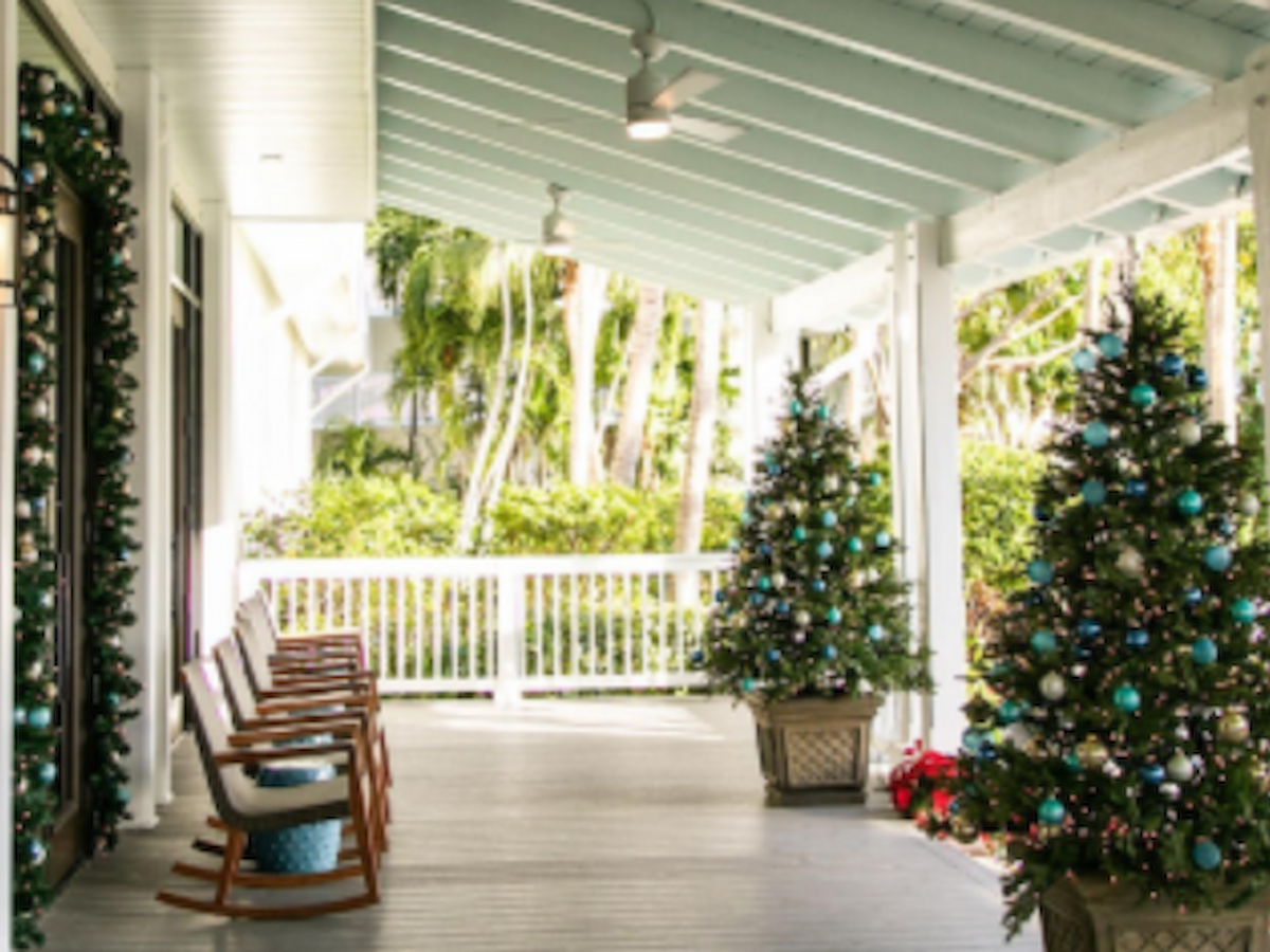 A porch with rocking chairs and decorated Christmas trees, surrounded by greenery and a light blue ceiling.
