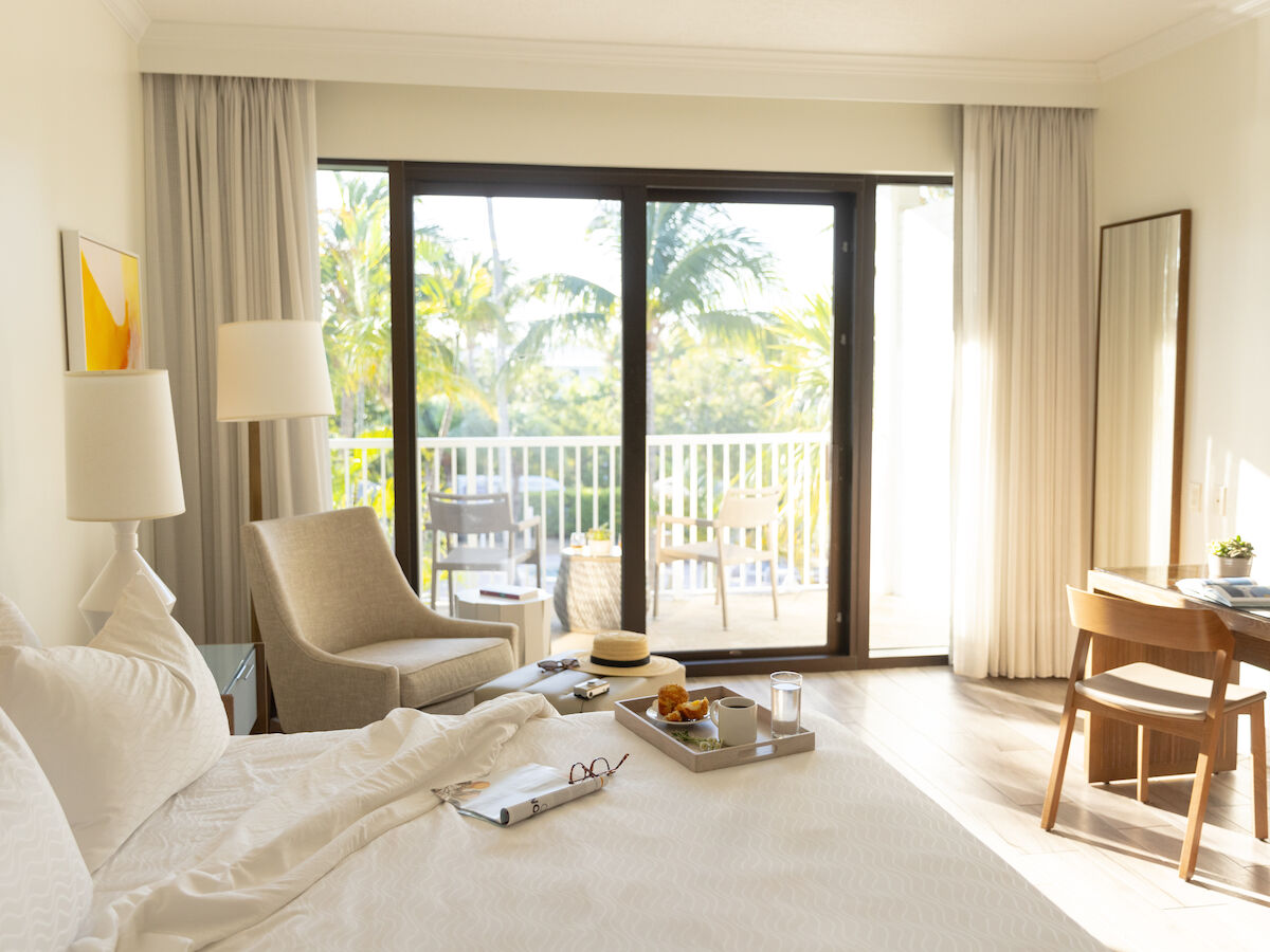 A bright hotel room with a bed, armchair, desk, and a sliding door leading to a balcony with a view of palm trees.