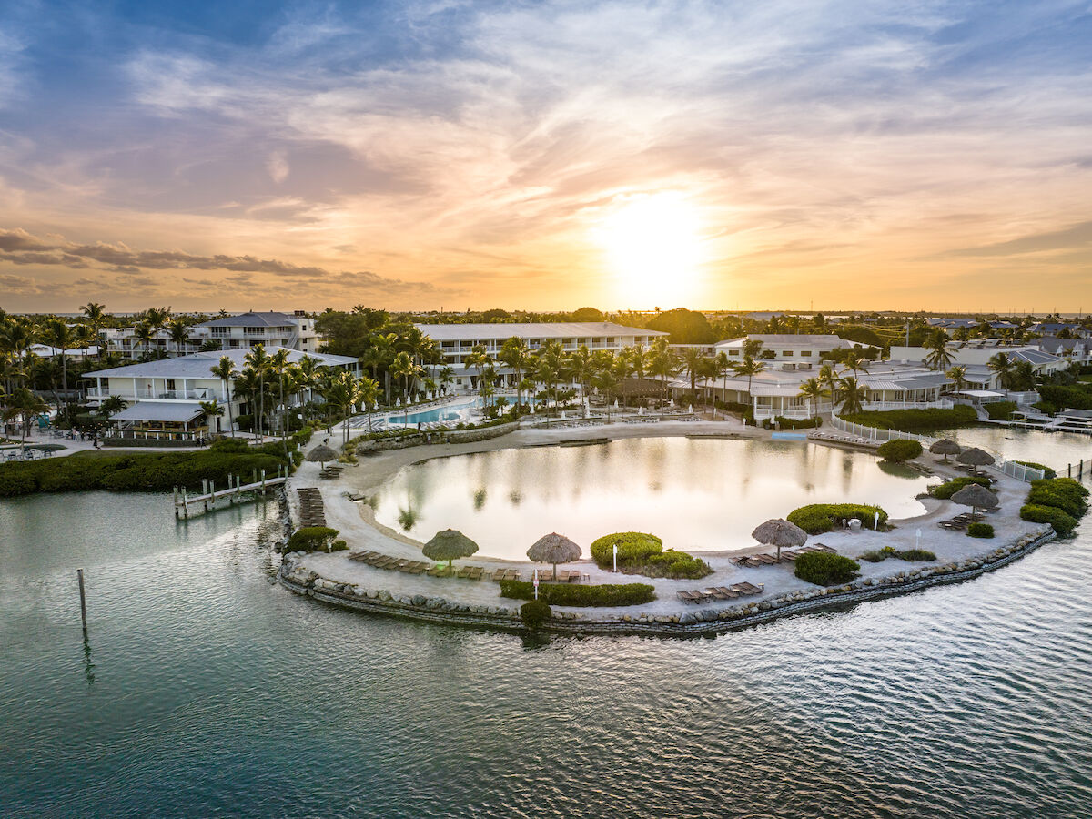 The image shows a beautiful coastal resort at sunset, featuring a lagoon, palm trees, and a sandy beach area with thatched umbrellas.