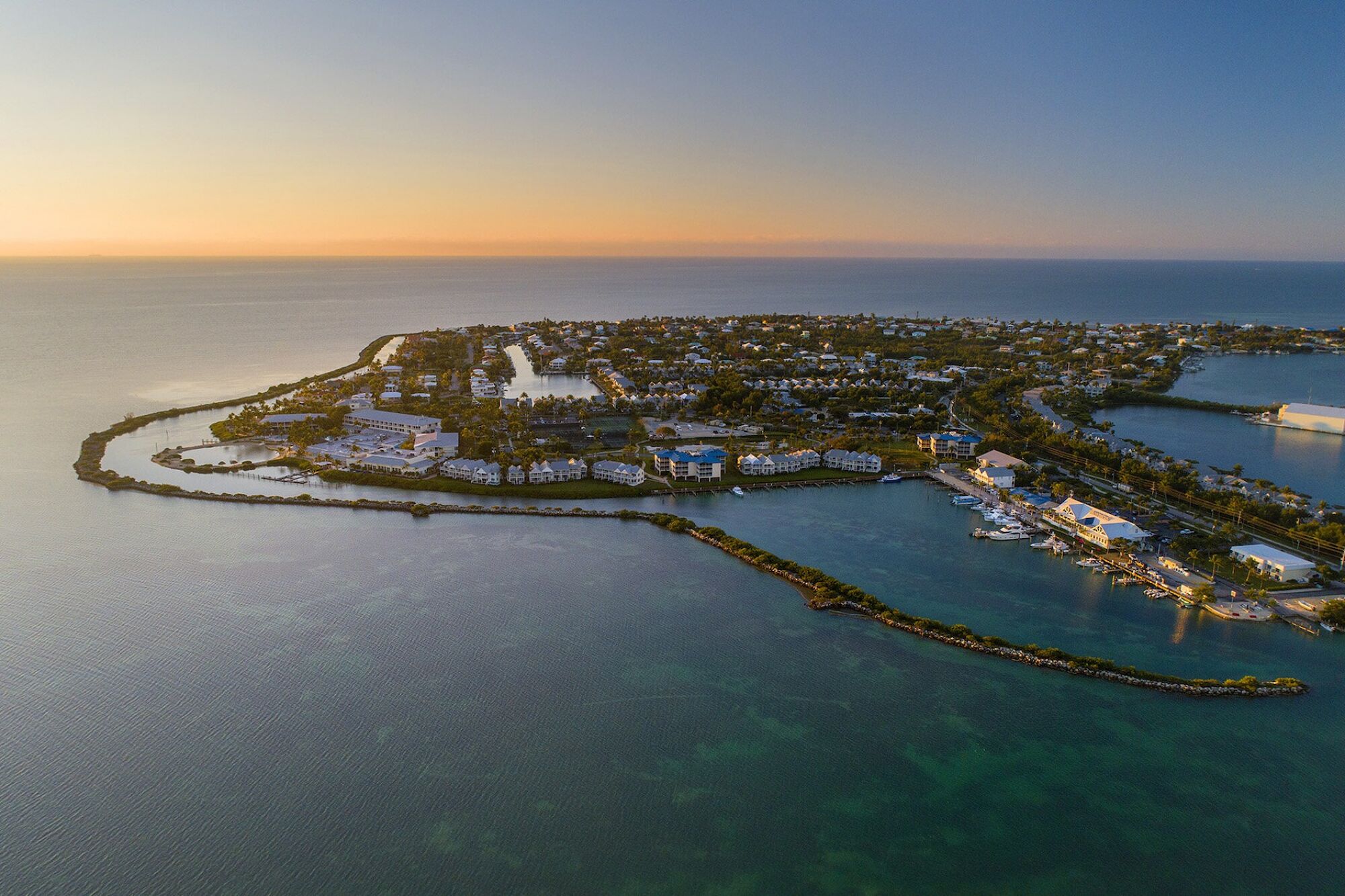 Aerial view of a coastal town on a peninsula with buildings, greenery, and surrounding ocean, under a sunset sky.