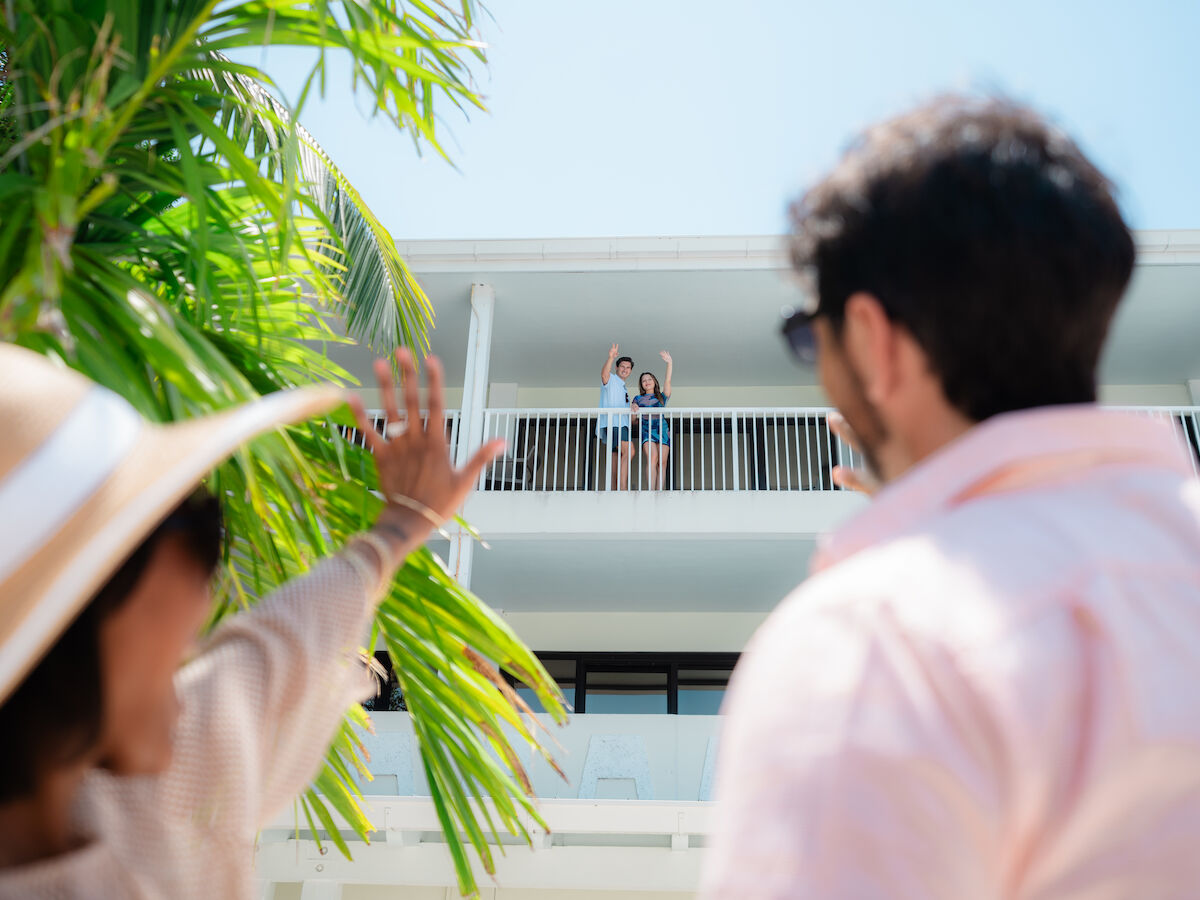 A couple waves from a balcony while two people on the ground, partially obscured by palm leaves, wave back.