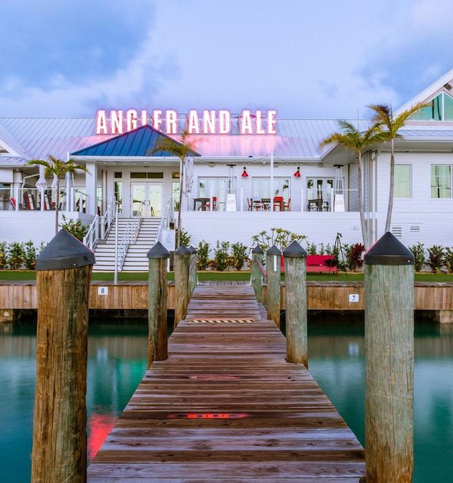 A waterfront restaurant named "Angler and Ale," with tropical landscaping, a wooden dock leading to it, and a serene body of water.