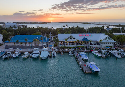 A marina with several boats docked, buildings by the water, and a sunset in the background.