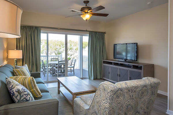 A living room with a sofa, decorative pillows, armchair, coffee table, TV, lamp, ceiling fan, and sliding doors leading to an outdoor deck.