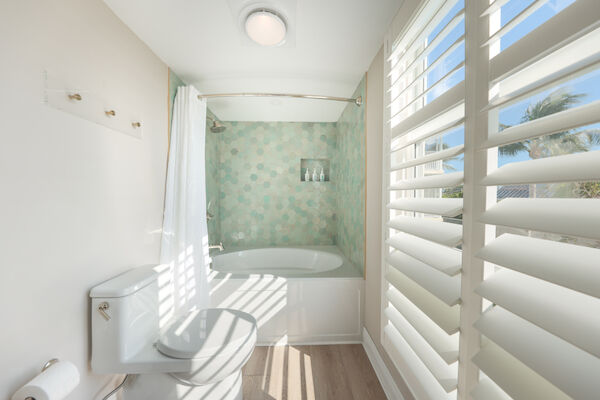 A small bathroom with a bathtub, hexagonal tiles, a toilet, and white shutters letting in sunlight.