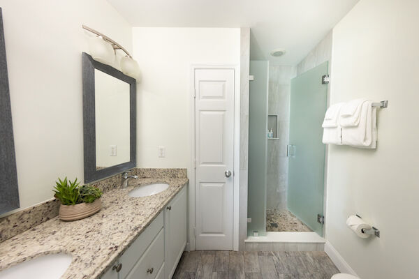 A modern bathroom with granite countertops, dual sinks, a glass shower, potted plant, towels on a rack, and a toilet with toilet paper.